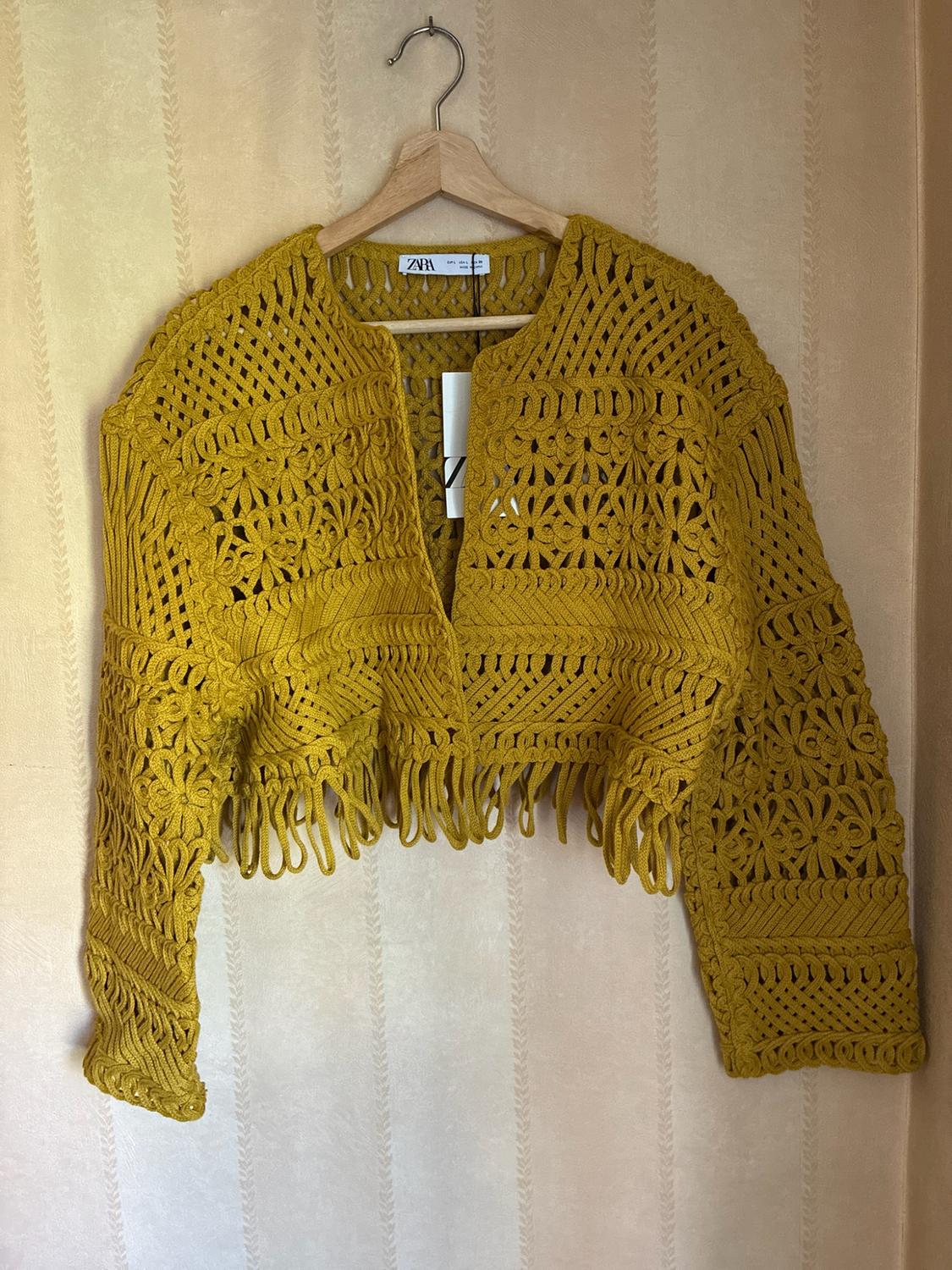 Short mustard open-knit jacket with fringing, hanging on a wooden hanger against a wallpapered wall. Label with the text ZARA.