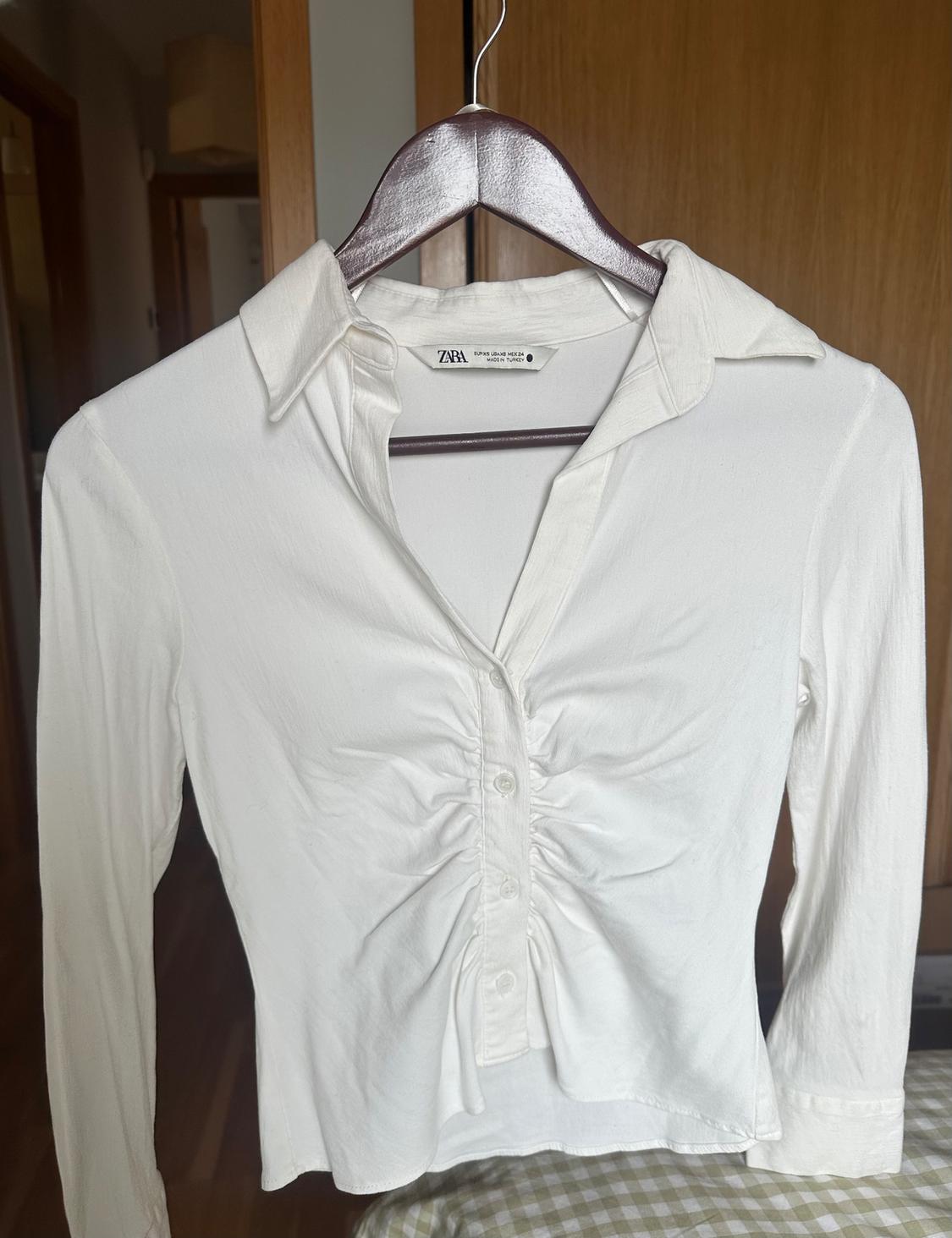 White long-sleeved shirt with ruching in the centre, hanging on a wooden hanger indoors. Visible tag: ZARA.