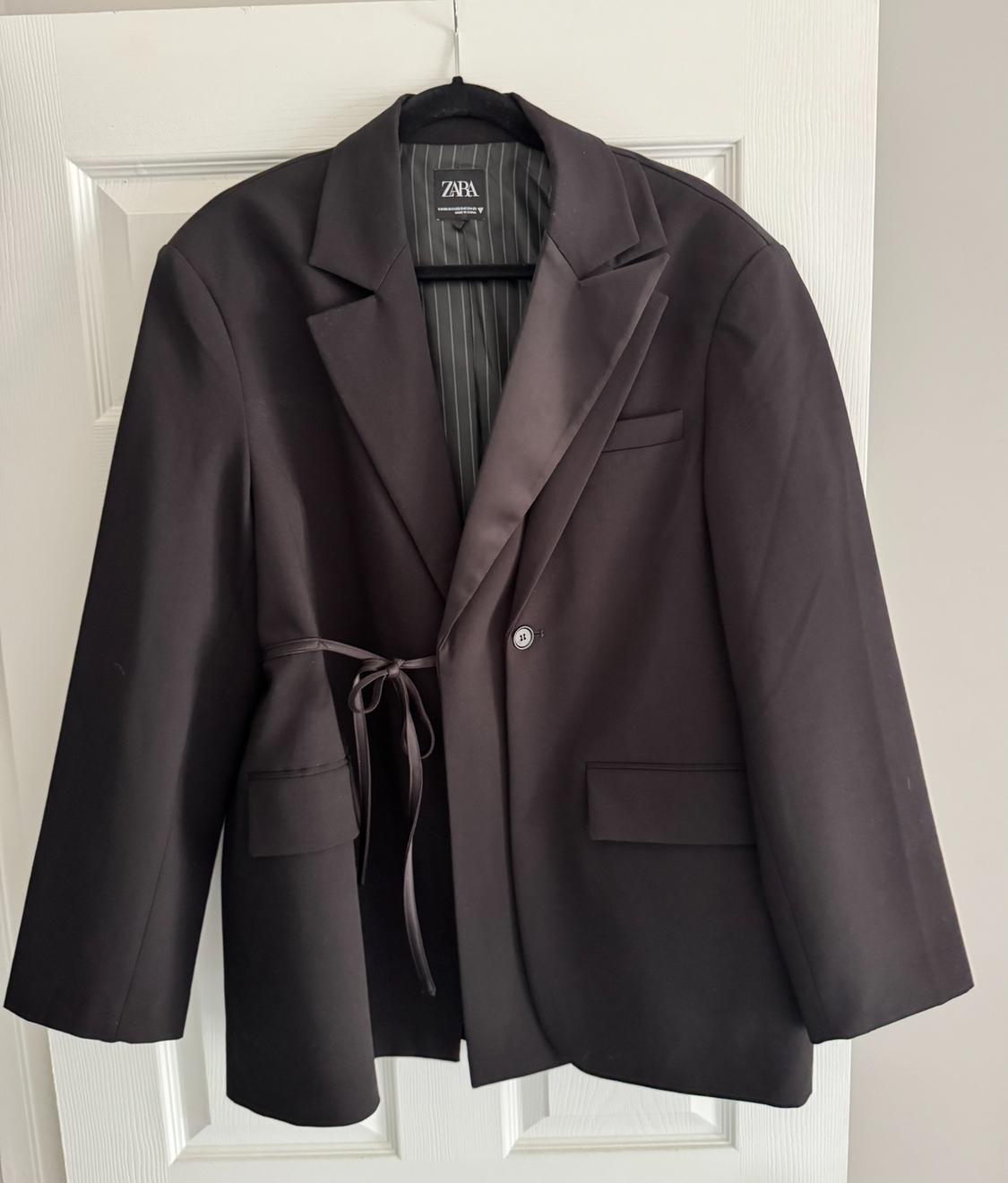 Black blazer hanging on a white door. Features a satin lapel, one button, bow detail, and striped lining. Label with the text ZARA.