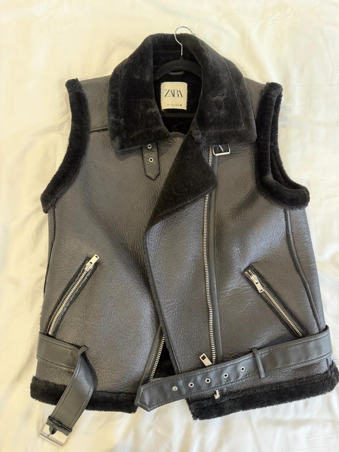 Black faux leather vest with fur lining, asymmetric zipper, pockets, and belt. ZARA label. Set against white fabric.