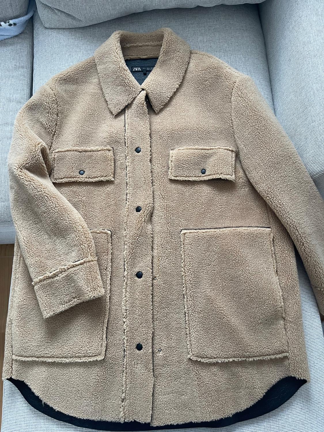 Beige wool jacket with black buttons and large pockets, placed on a grey sofa. Label visible with text "ZARA".