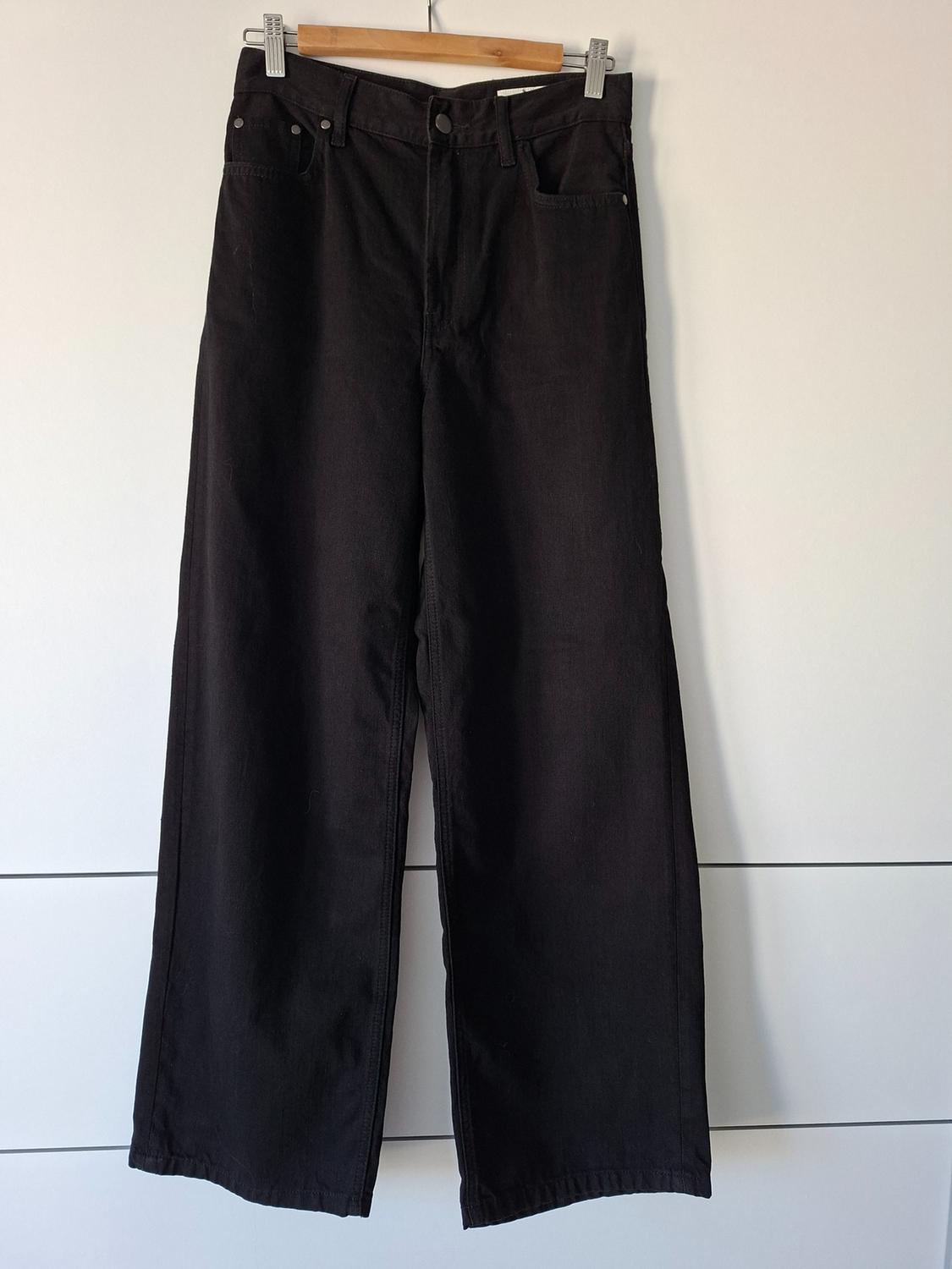 Black wide-leg jeans hanging from a wooden hanger against a white background with horizontal lines.