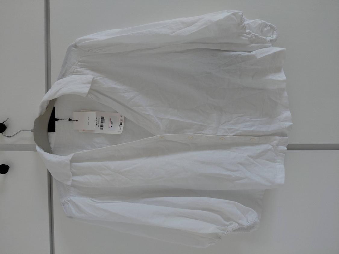 White long-sleeved shirt with a collar, buttons and elasticated cuffs, hanging on a hanger against a white background. Label visible.