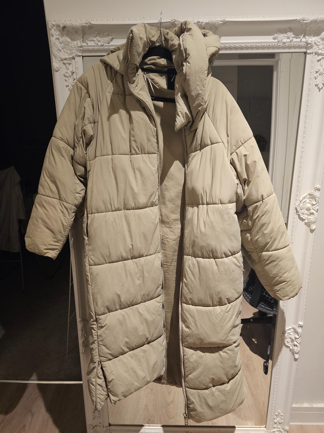 Beige padded coat hanging on a decorative white-framed mirror. Partial reflection of a room in the background.