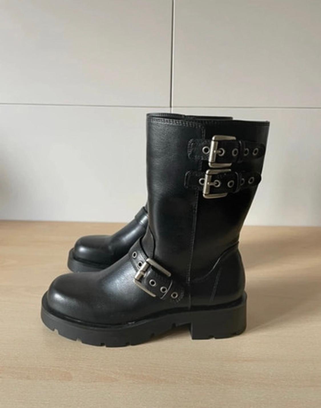 Black leather boots with a chunky sole and metallic buckles on the side, against a white wall and light wooden floor background.