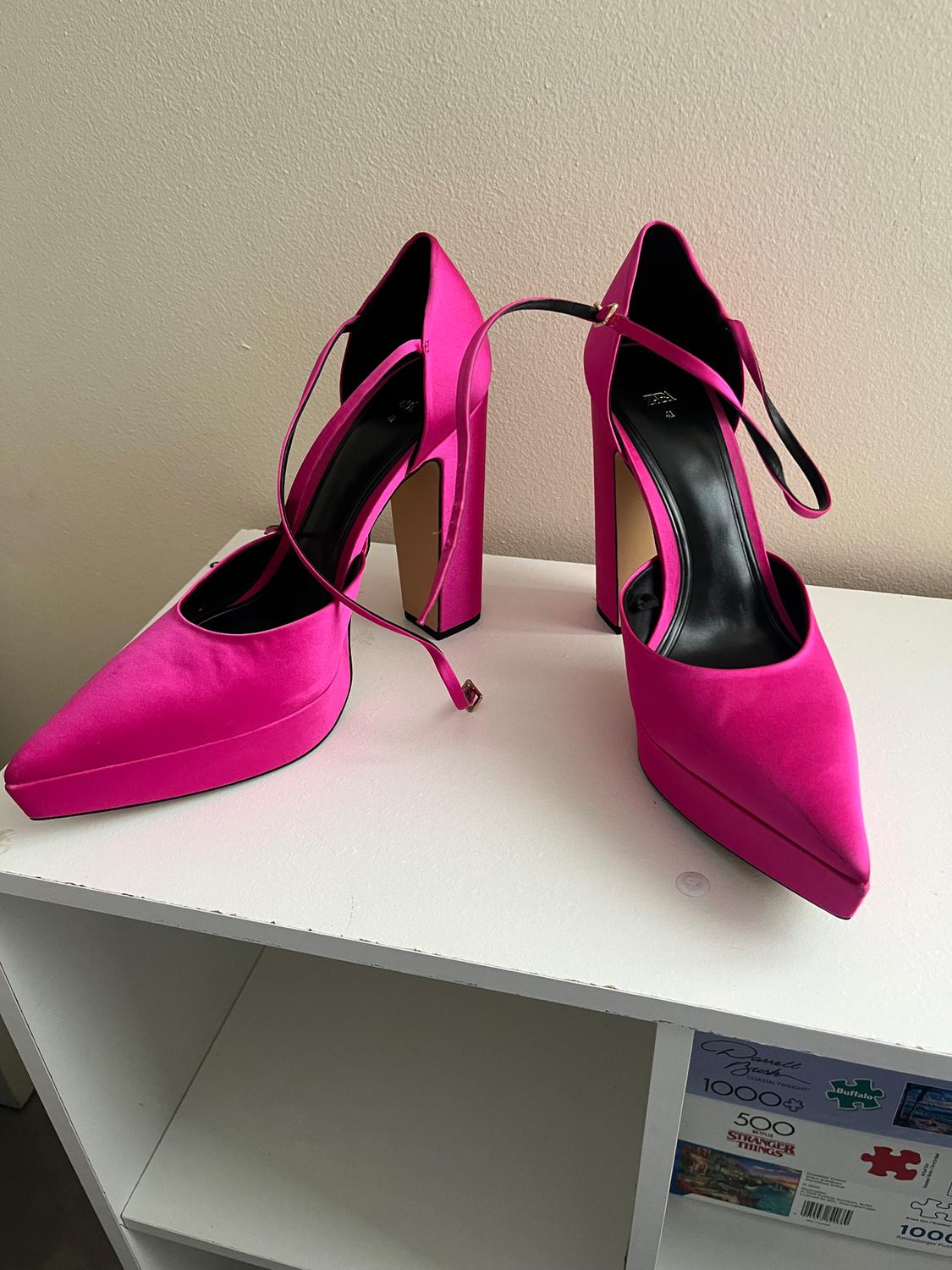 Fuchsia high heels with thin straps on a white shelf. Beige wall background. Jigsaw puzzle box visible below.