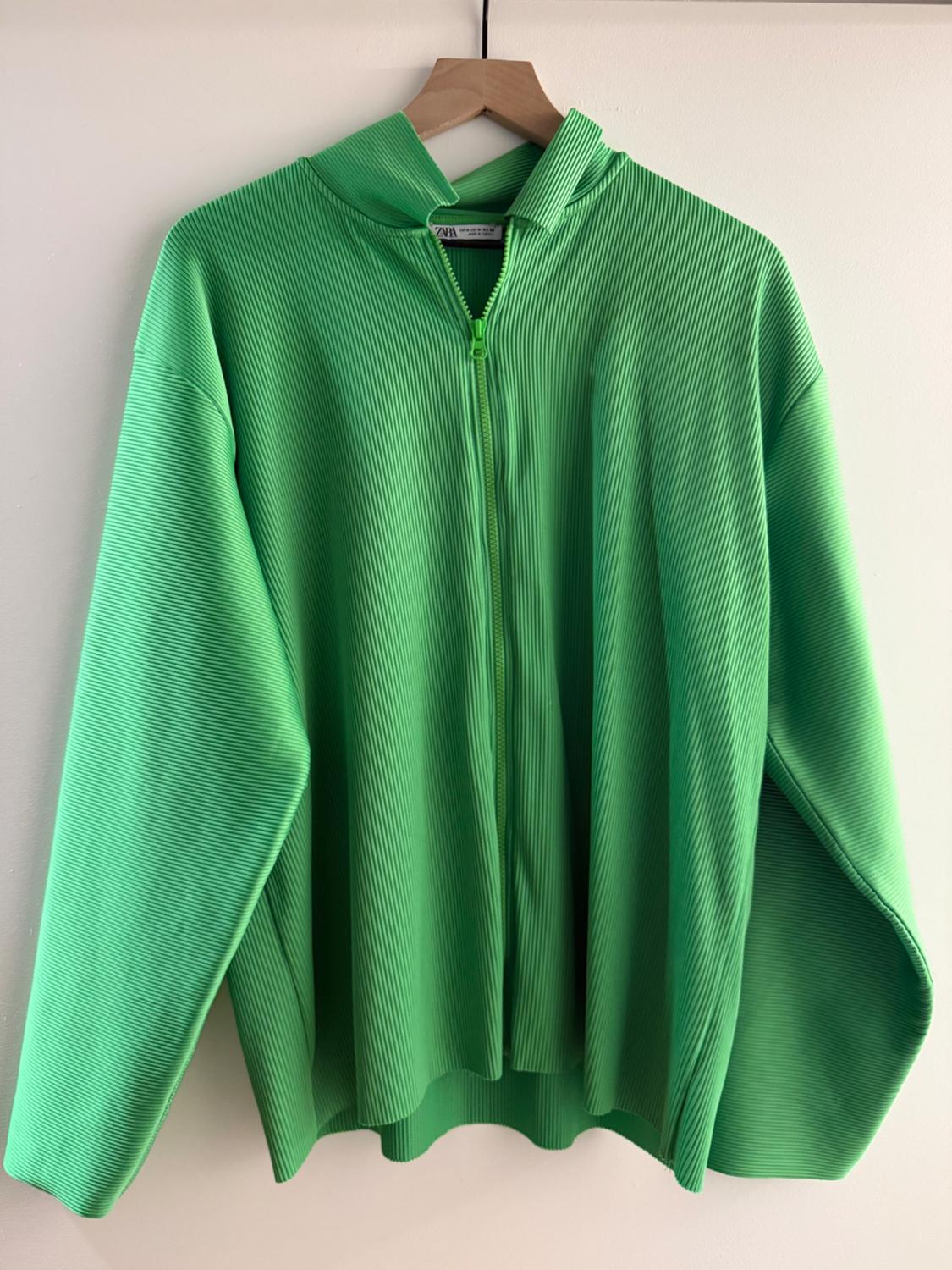 Green ribbed fabric jacket hanging on a wooden hanger. It has a front zip and a hood. White background.