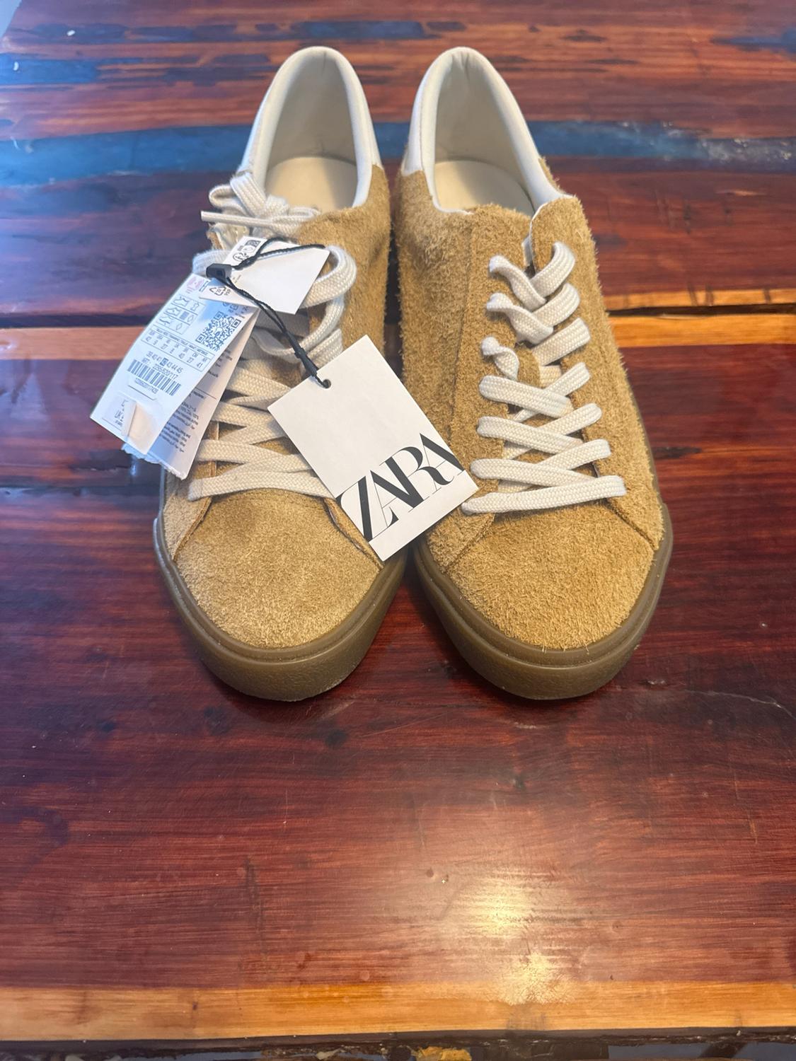 Mustard suede trainers, white laces and brown sole on a wooden table. Label with the text ZARA.