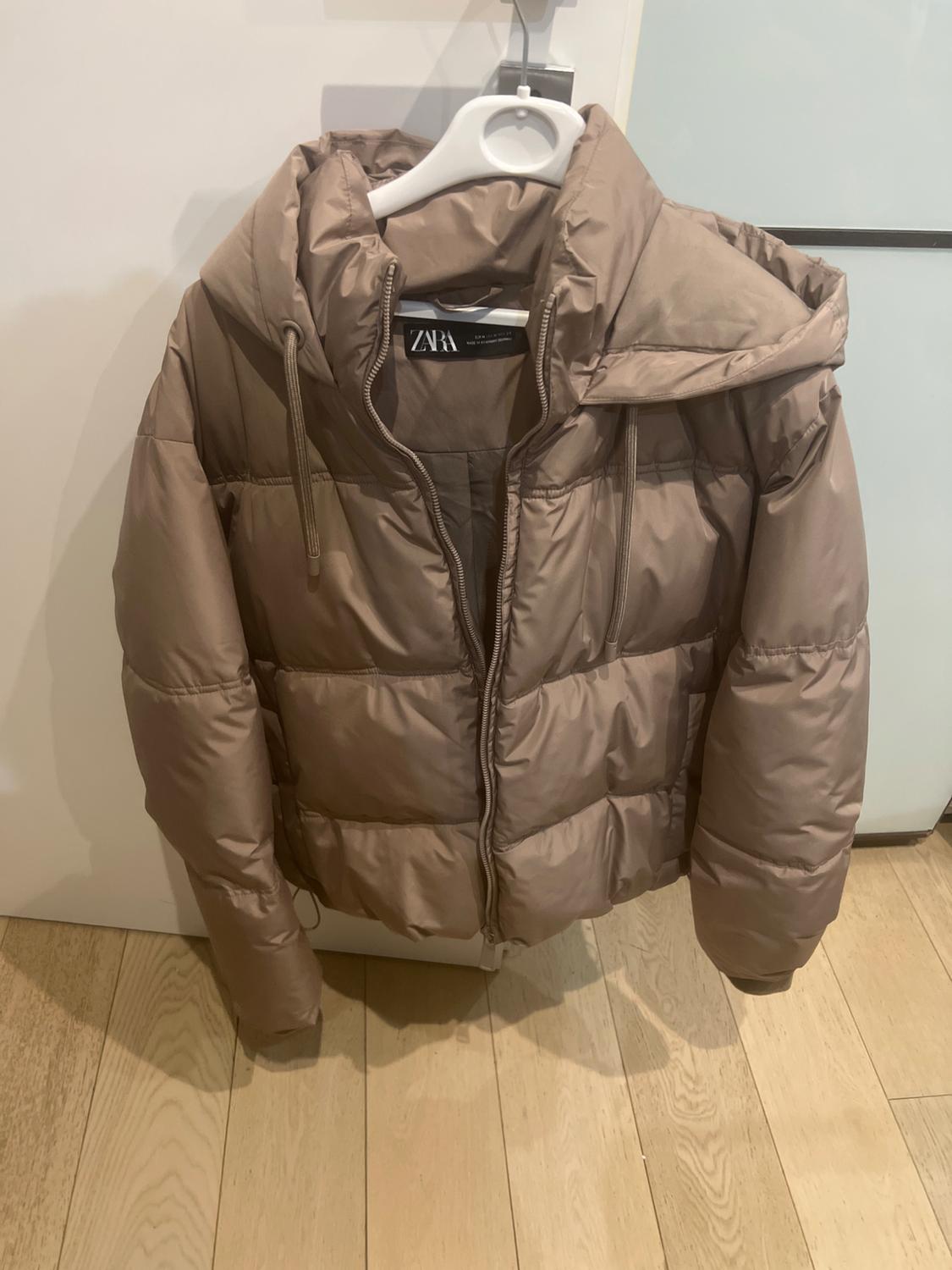 Light brown padded jacket hanging on a white hanger over a light wooden floor. Visible label with text "ZARA".