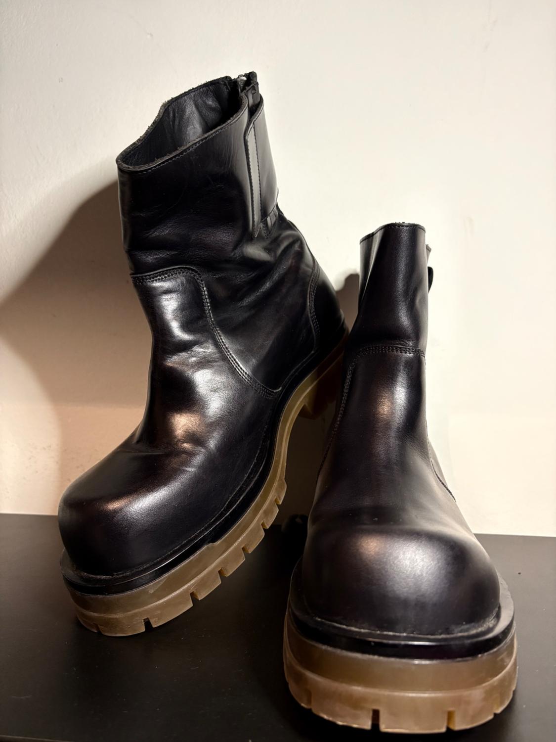 Black leather boots with a chunky brown sole, placed on a dark surface, illuminated by soft light.