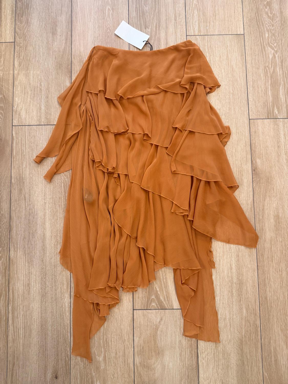 Brown skirt with cascading frills and an asymmetrical hem, laid out on a wooden floor. It has a white label at the waist.