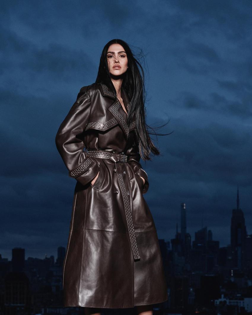 ZARA x Ludovic de Saint Sernin: Kolekcija koja definira novu estetiku senzualnosti 2 Woman in a long, dark, studded coat, standing in a nighttime urban landscape with a cloudy sky and buildings in the background.