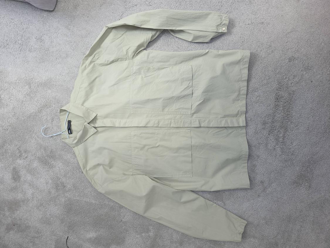 Long-sleeved beige shirt with a classic collar, placed on a grey rug. Visible label on the collar.