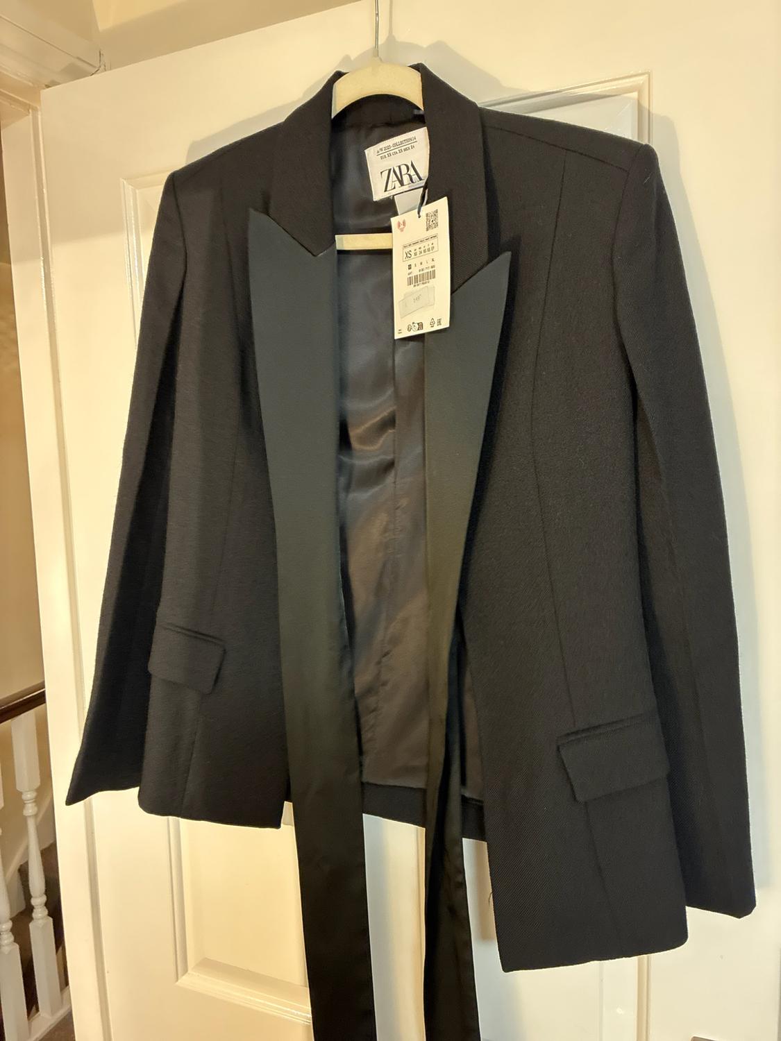 Black blazer with satin lapels and cape-style sleeves, hanging on a white door. Interior label with the text ZARA.