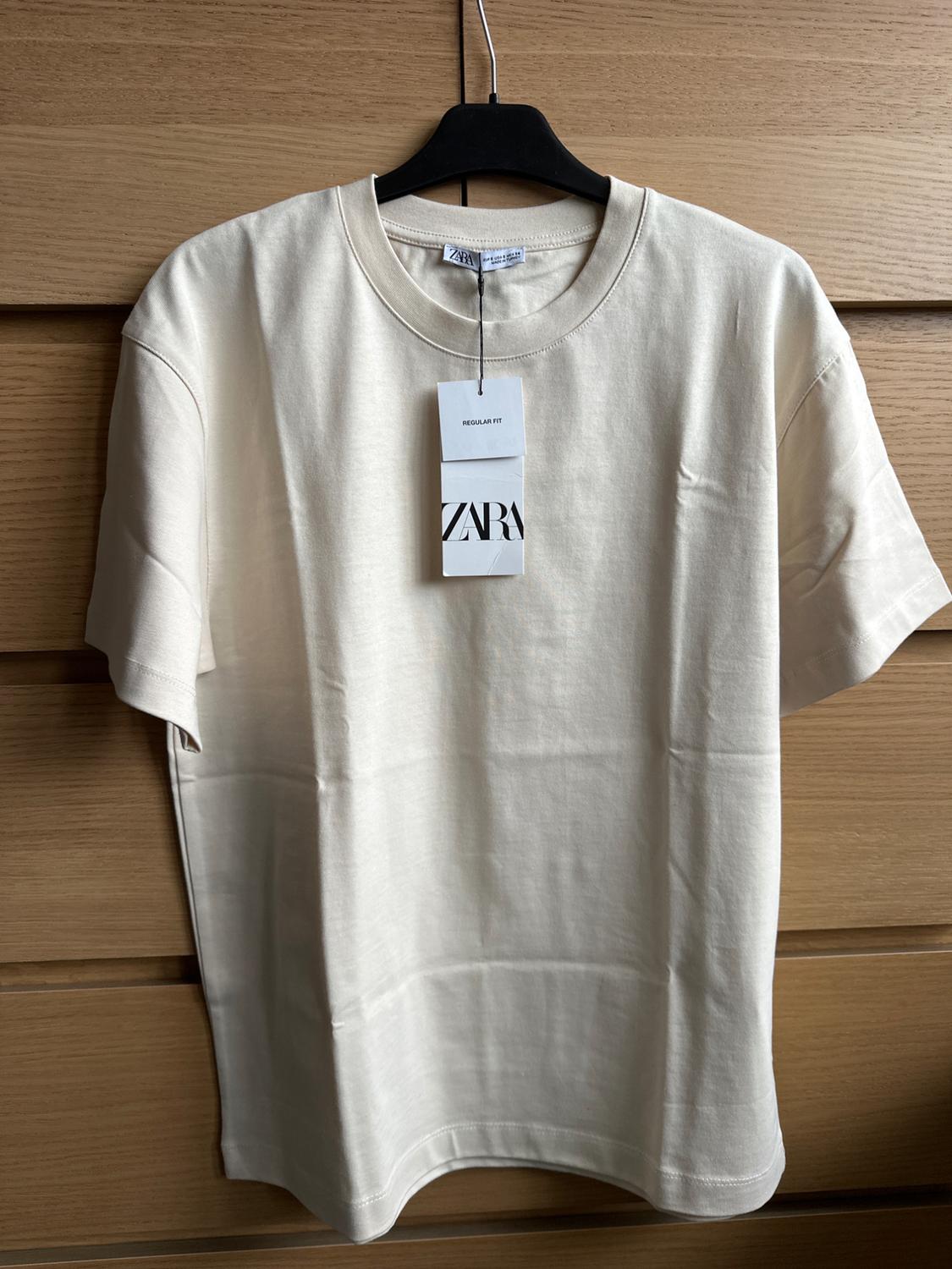 White short-sleeved T-shirt hanging on a black hanger against a wooden background. Visible label with text "REGULAR FIT" and "ZARA".