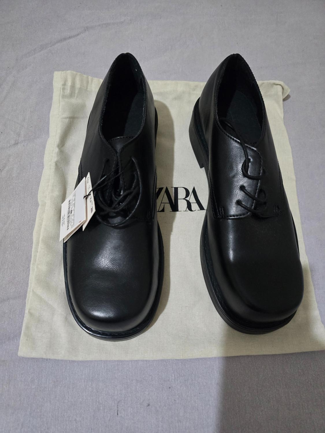 Pair of black lace-up shoes with tags on a fabric bag bearing the text ZARA, placed on a grey surface.