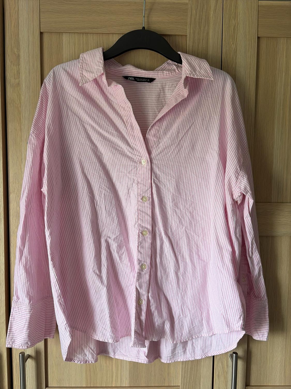 Pink and white striped button-down shirt hanging on a black hanger in front of a wooden wardrobe. The label reads ZARA.