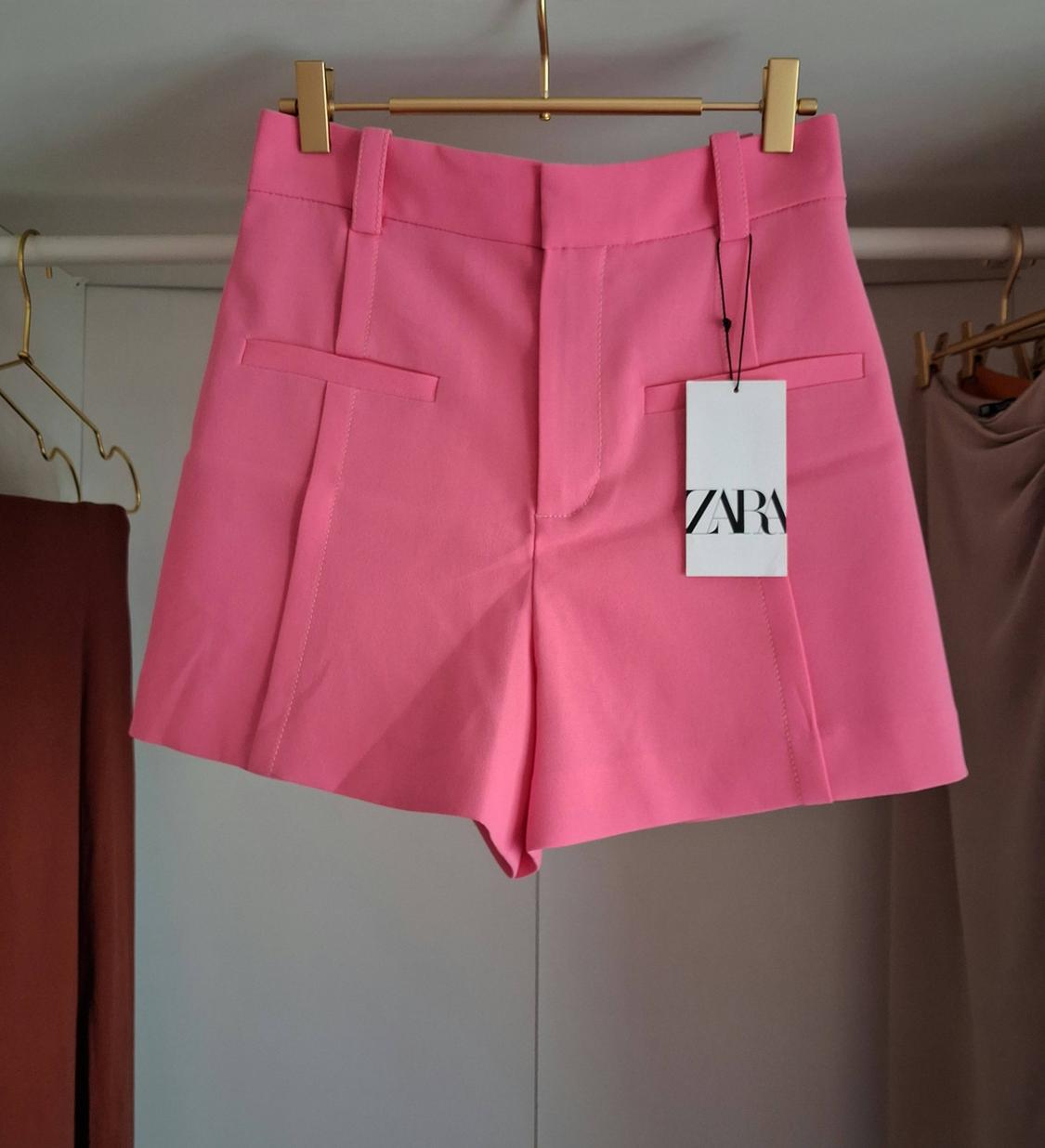 Pink shorts hanging on a gold hanger in a wardrobe next to other garments. Tag with the text ZARA.
