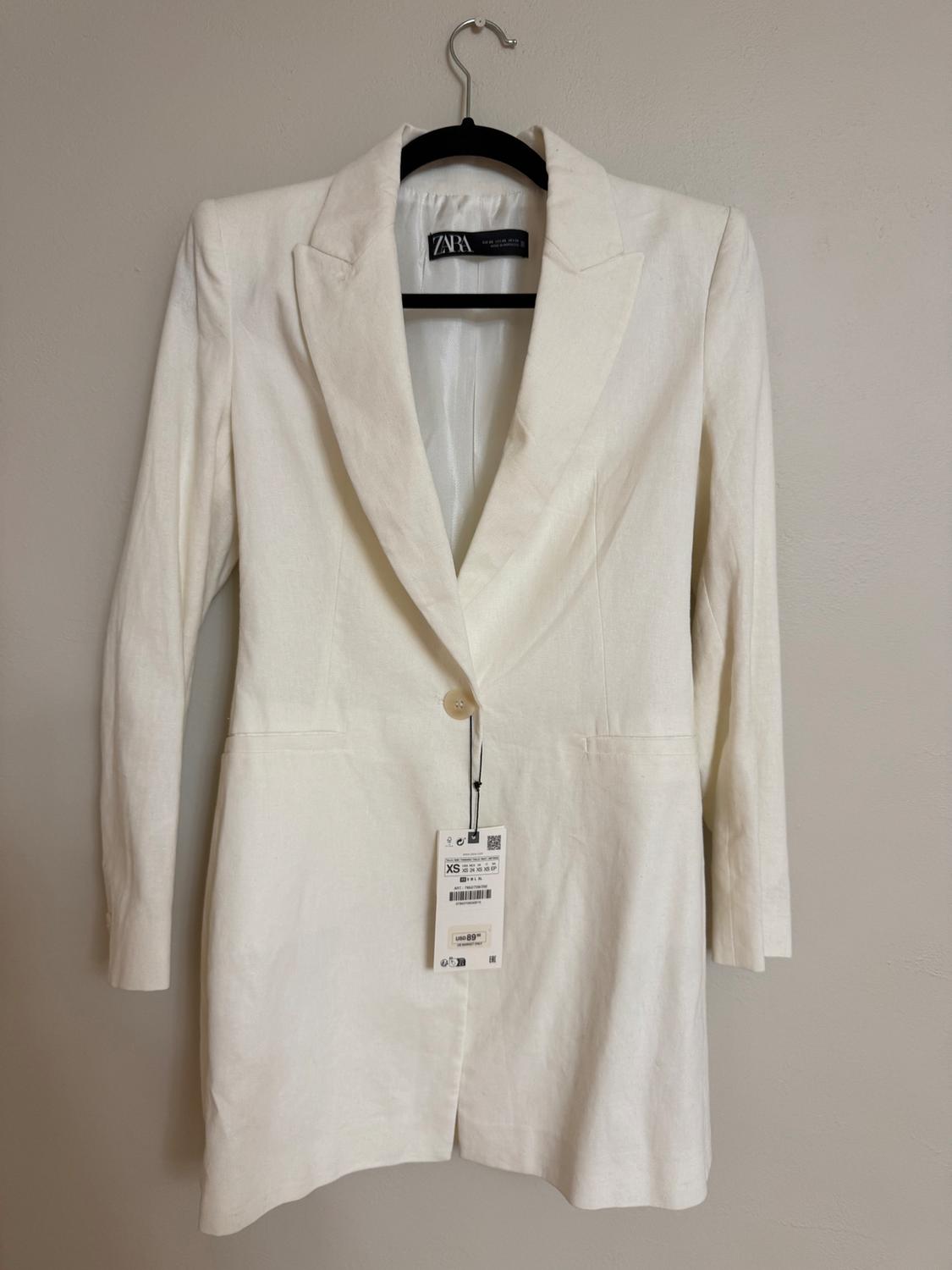 Long-sleeved white blazer hanging on a black hanger. Visible tag with XS size details and barcode. Zara brand.