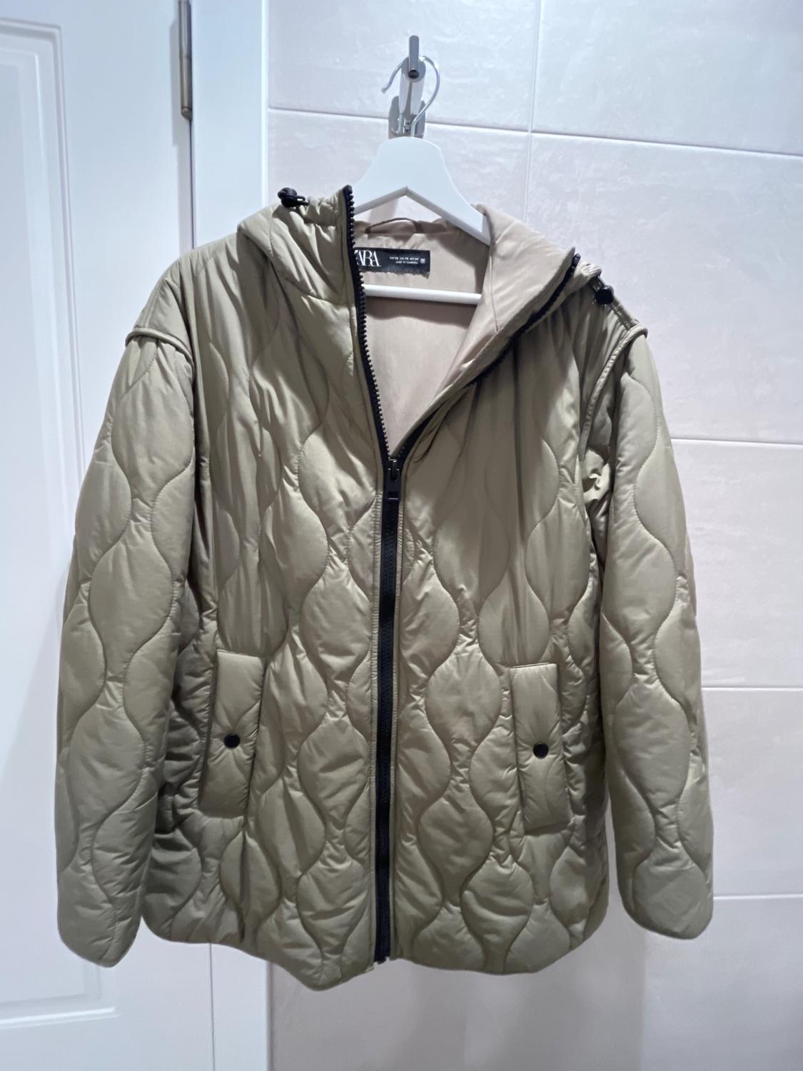 Beige padded jacket with a hood and black zip, hanging on a white hanger against a white tiled wall.