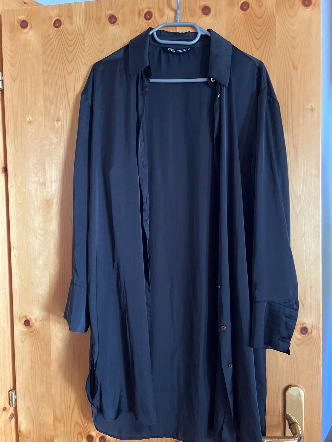 Dark, long-sleeved, unbuttoned shirt on a hanger on a wooden door. The label on the collar shows the text ZARA.
