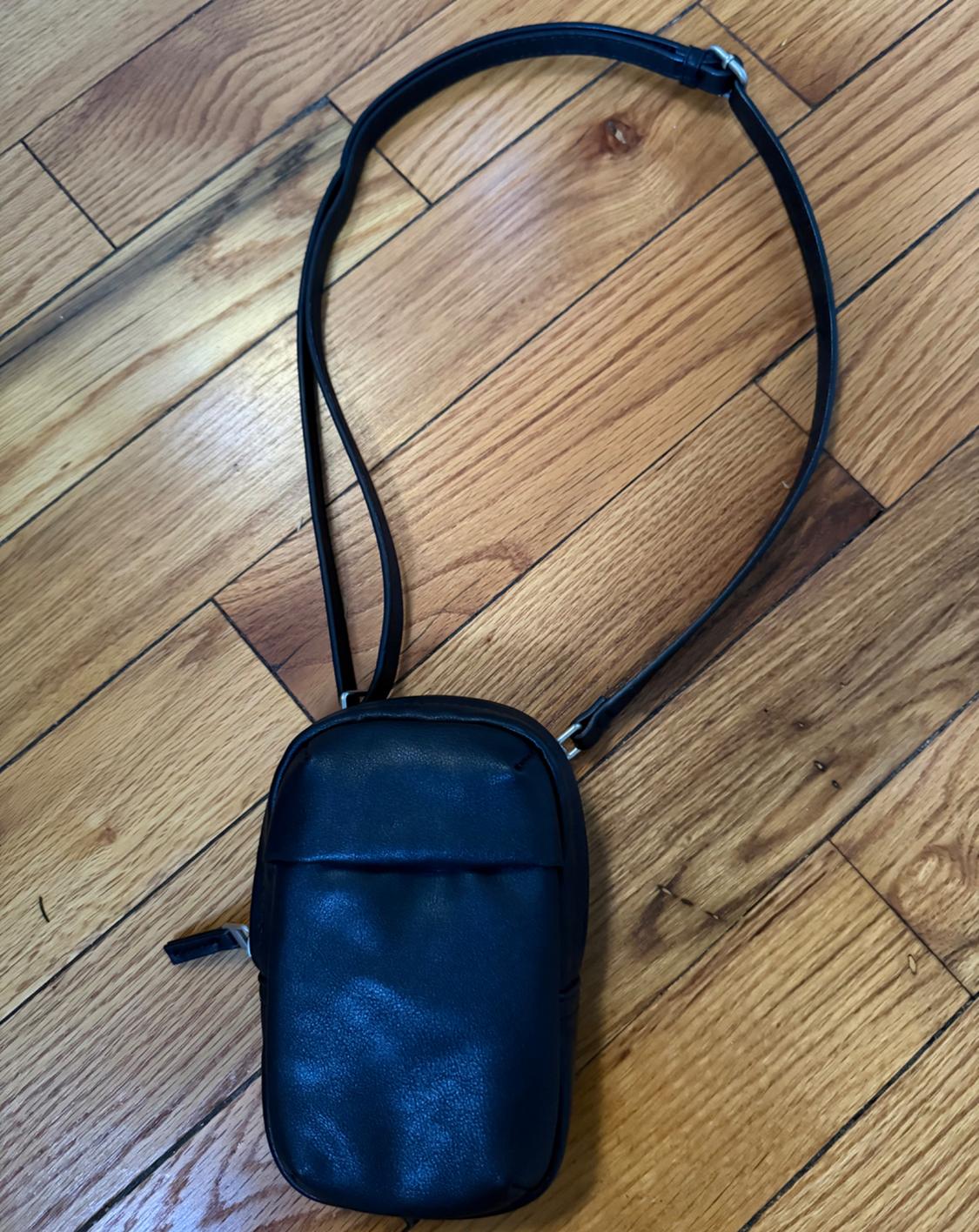A small black crossbody bag with a thin strap, placed on a light brown wooden floor.