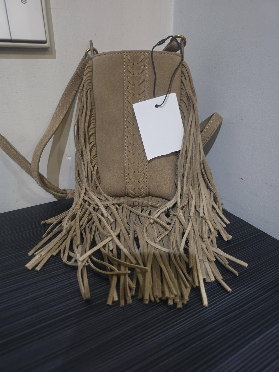 Brown suede crossbody bag with long fringing, braided detail and white label, on a dark surface against a wall.
