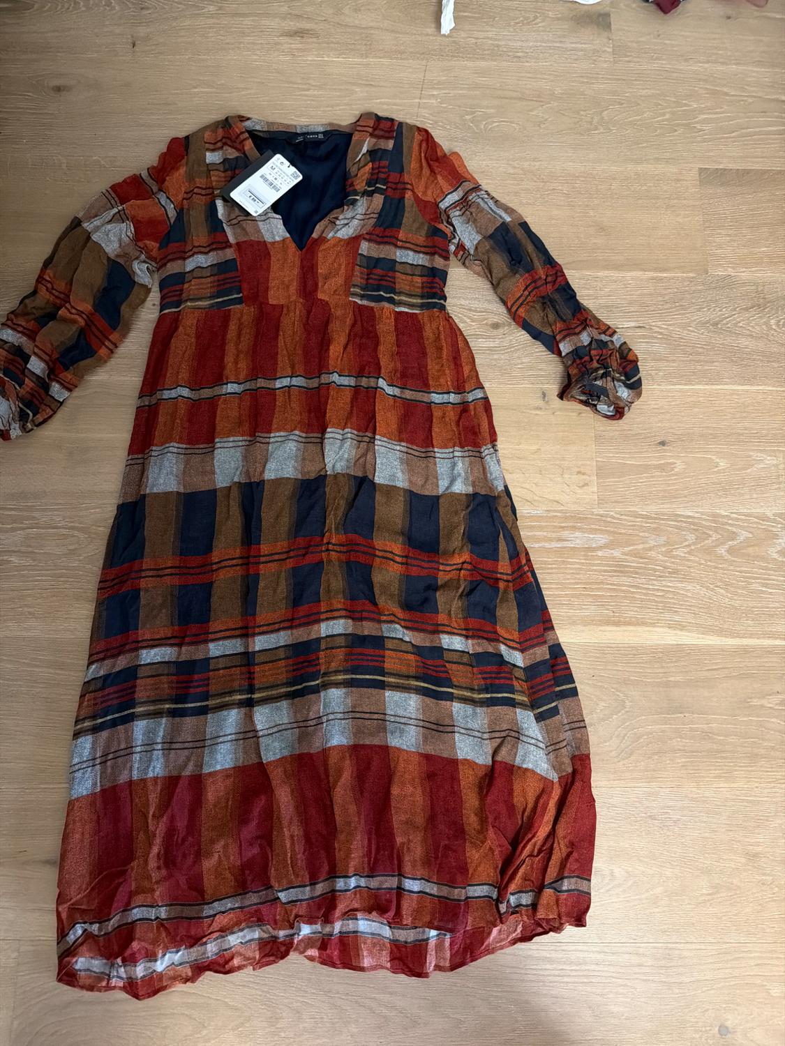 Checked dress in red, brown, and blue tones, with long puff sleeves, on a wooden floor. Visible label on the collar.