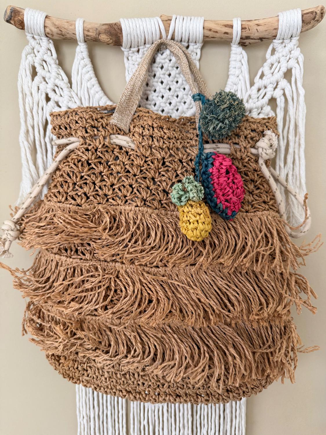Straw woven bag with fringe hanging on a wall, decorated with colorful pom-poms. White macrame background.