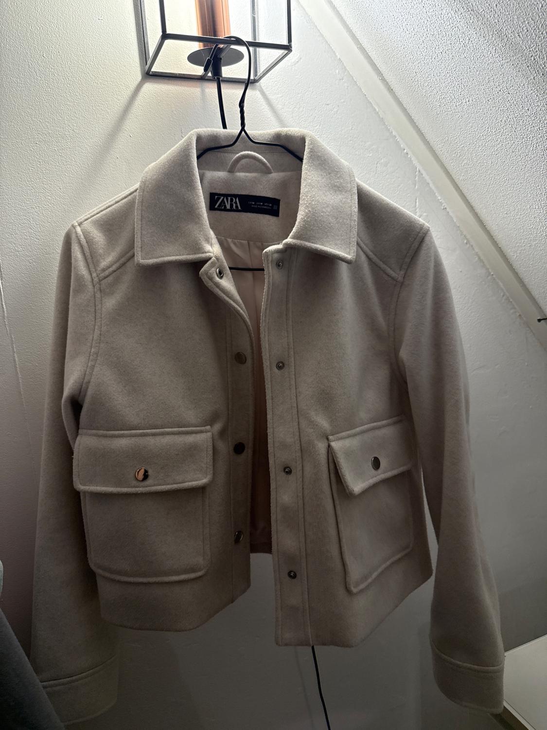 Beige wool jacket hanging on a hanger under a lamp. Visible label with the text "ZARA". Large pockets on the front.