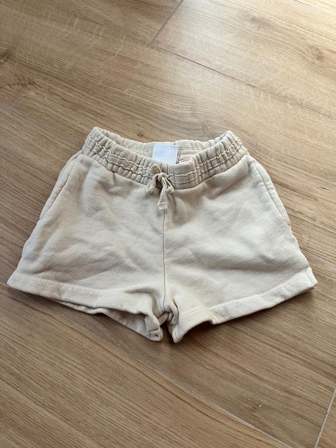 Beige shorts with elasticated waistband and drawstring, on light wood flooring. Label visible on the inside.