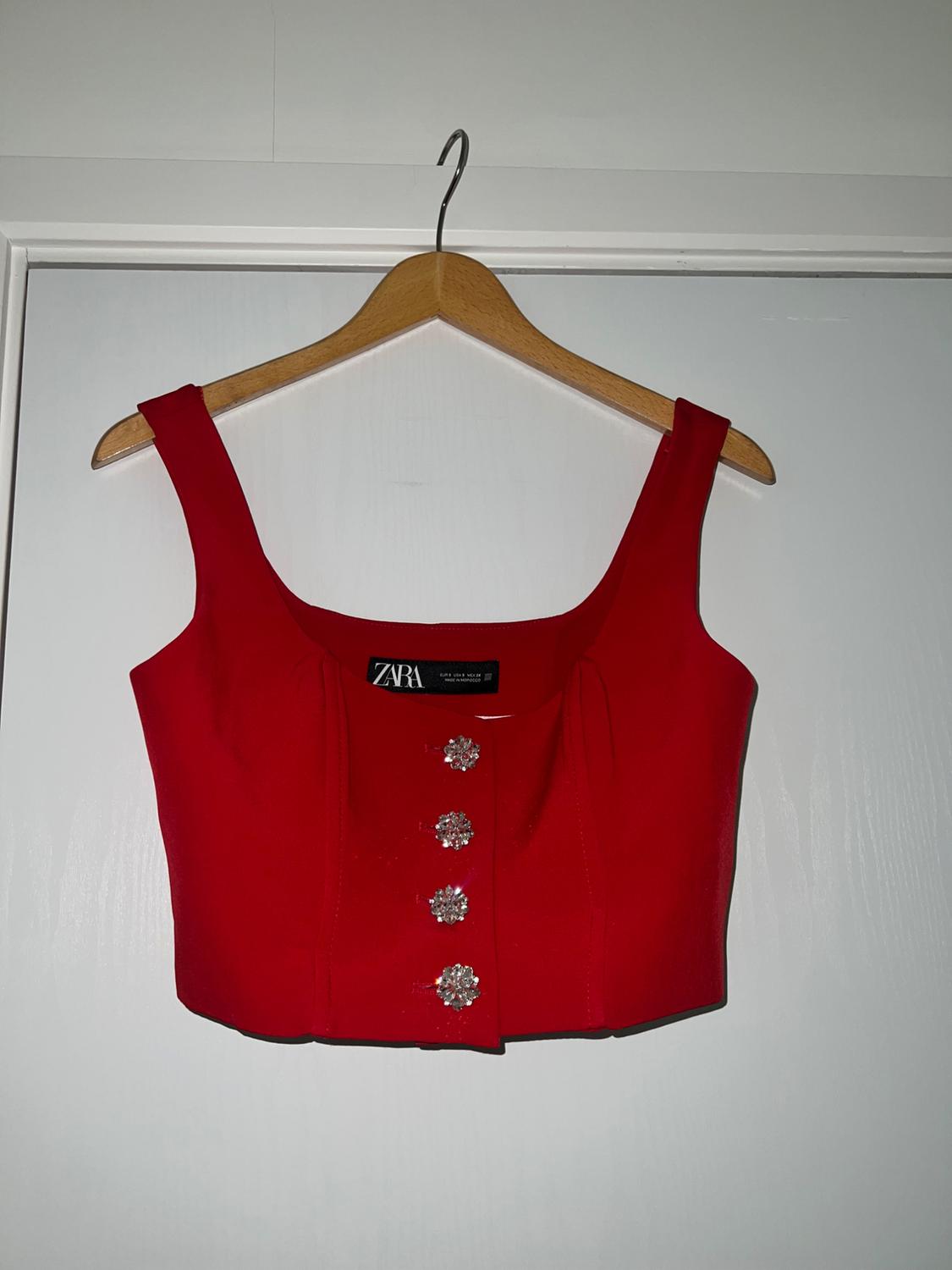 Red sleeveless top on wooden hanger on white door. Features a square neckline, four jewel buttons on the front and a ZARA label.