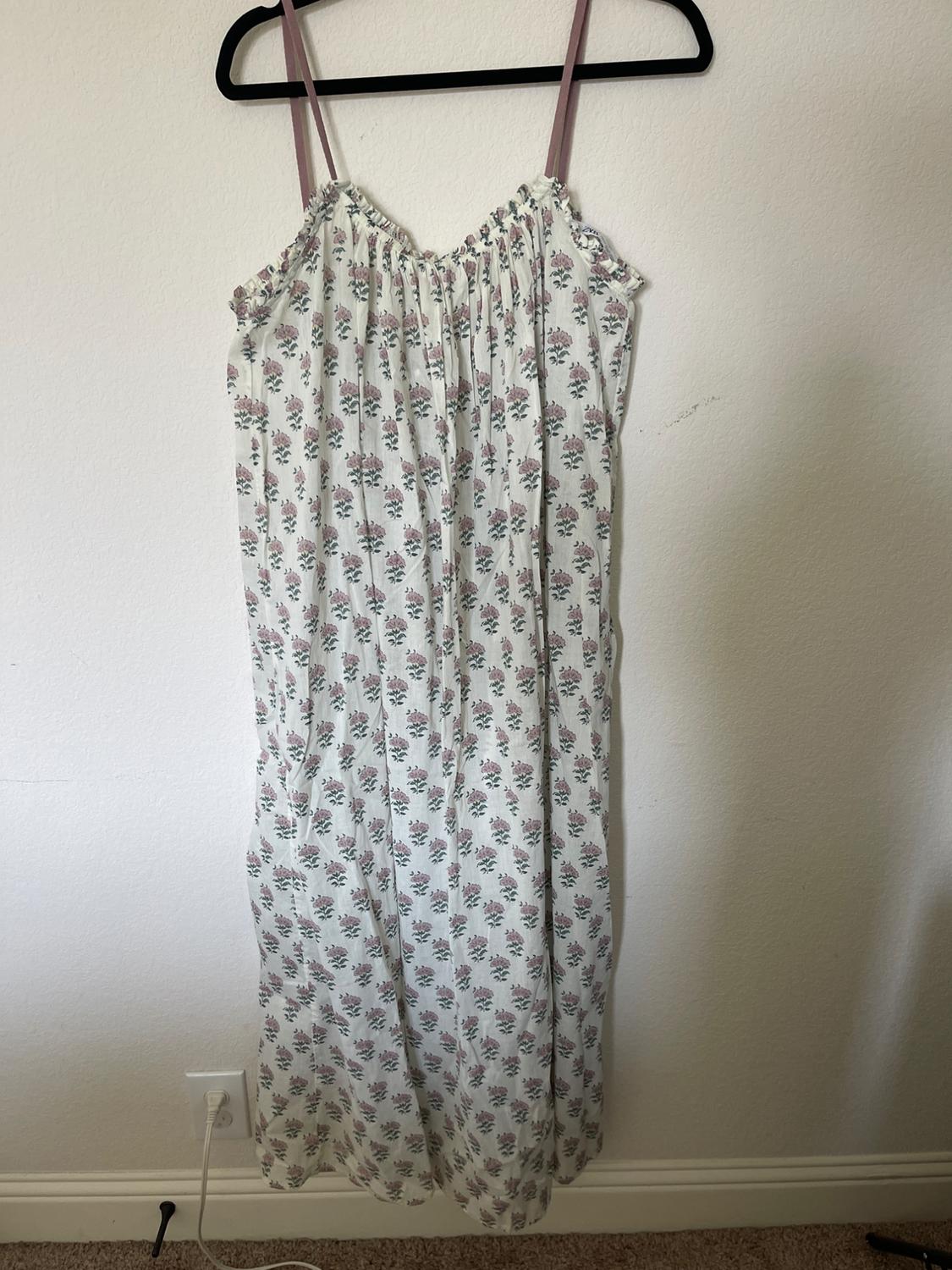 Long dress with thin straps and a pastel-toned floral print, hanging on a black hanger against a white wall.