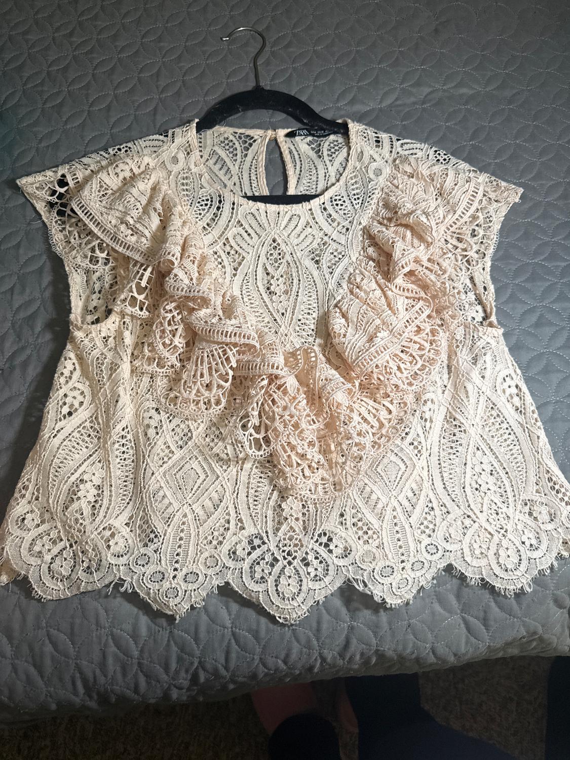Beige lace blouse with ruffles, hanging on a black hanger over a gray comforter with a circular pattern.