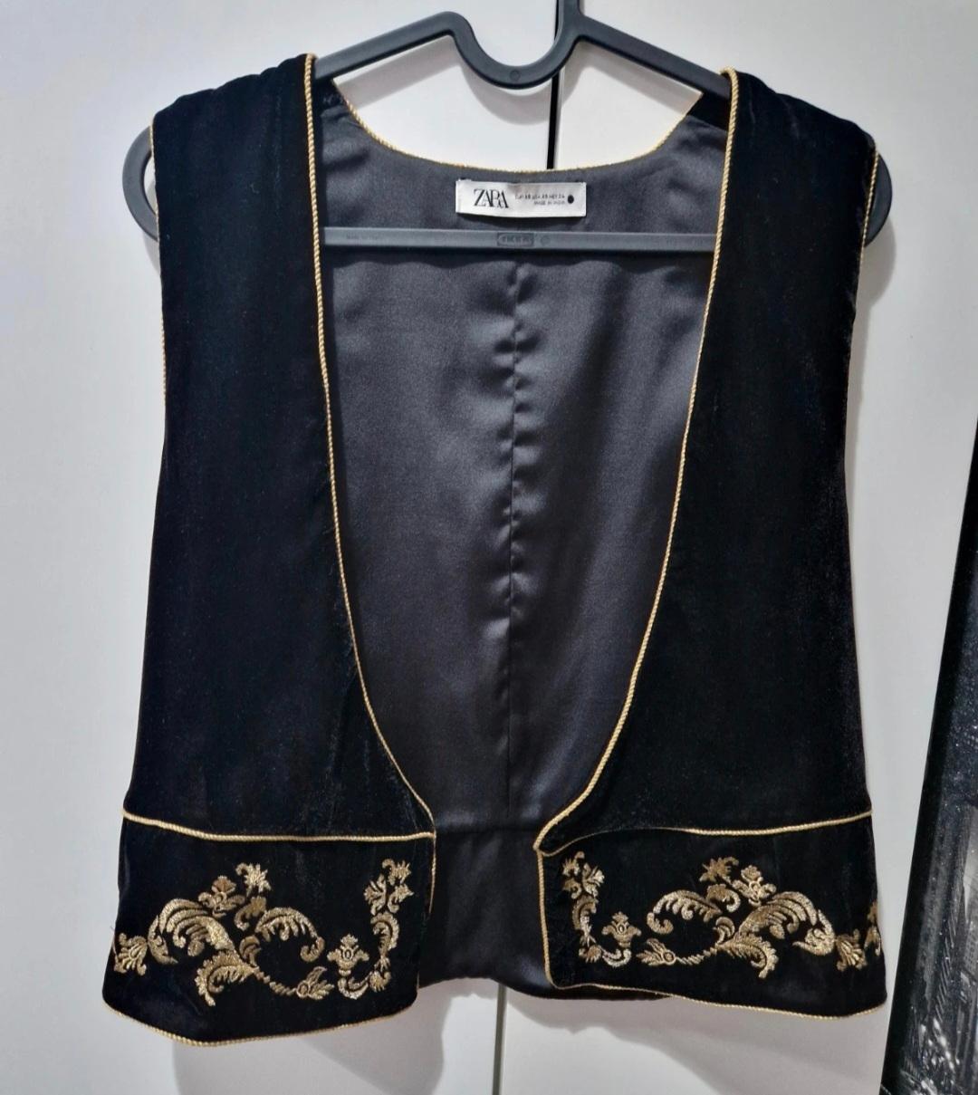 Black velvet waistcoat with gold embroidery on the pockets and gold trim, hanging on a grey hanger. Visible label.