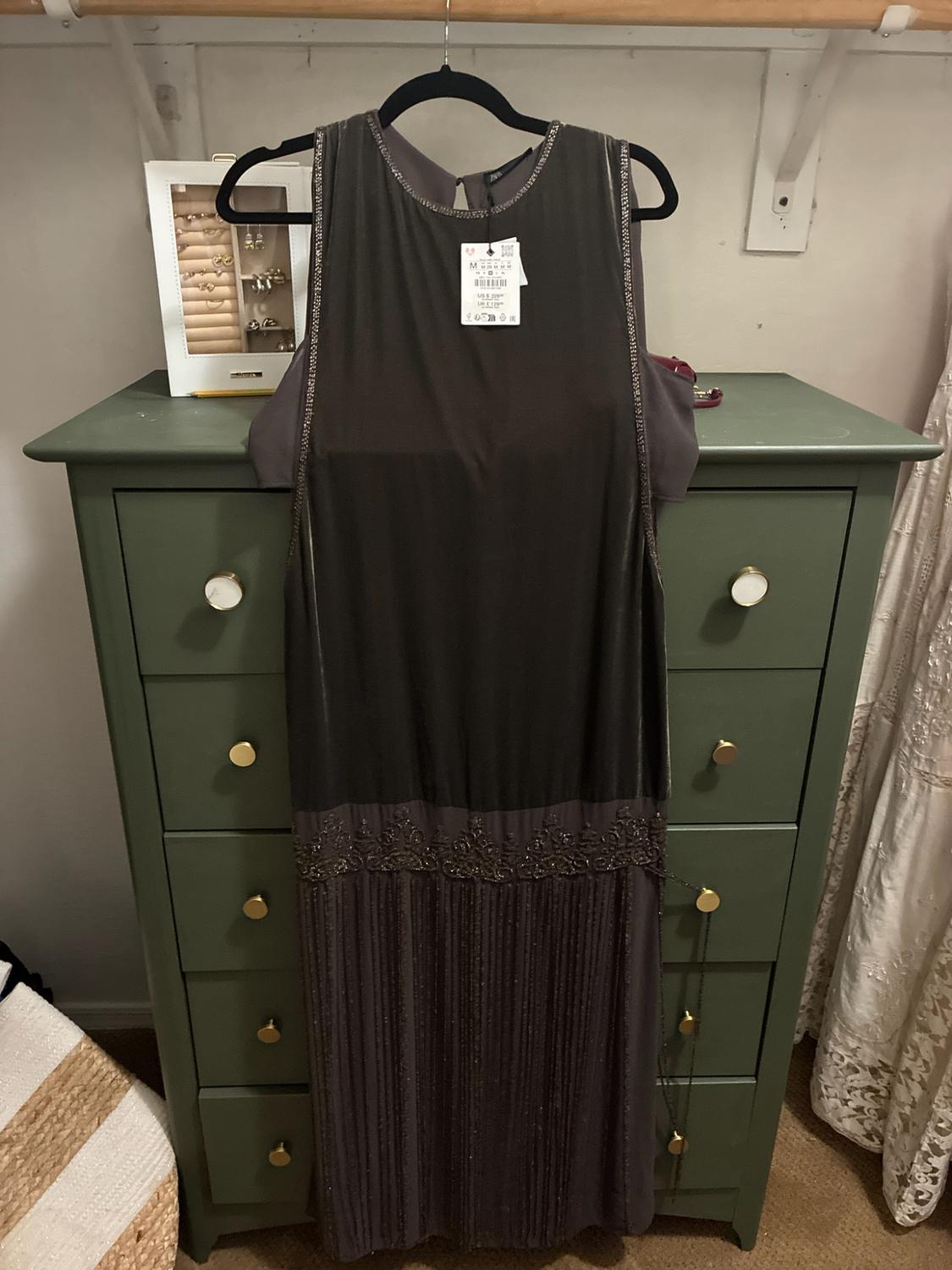Dark gray dress with beading, fringe skirt and tags, hanging on a hanger in front of an olive green dresser.