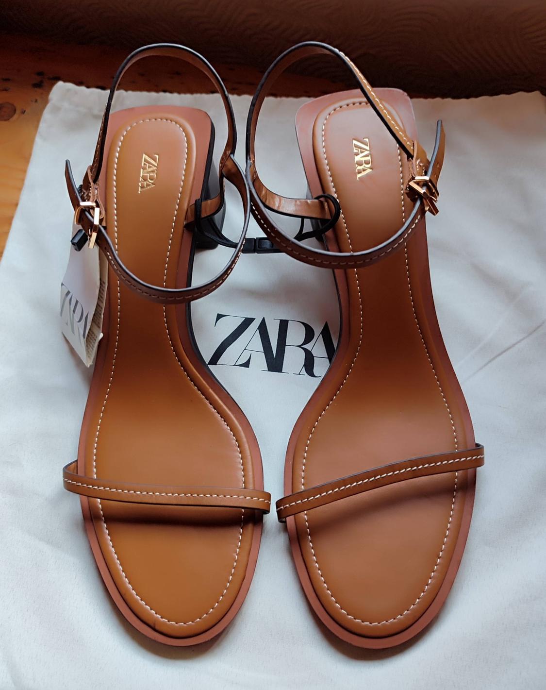 Brown heeled sandals with straps on a white bag. The text “ZARA” is visible on the insoles, the label and the bag.