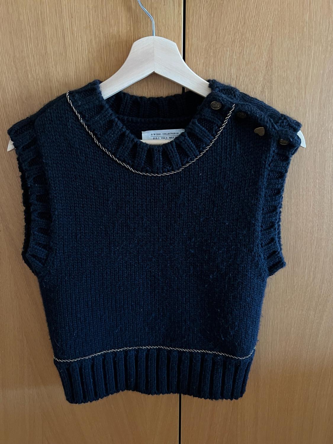 Navy blue knitted waistcoat with buttons and gold chain on a hanger against a wooden background. Text: F/W 2018 - COLLECTION 04.