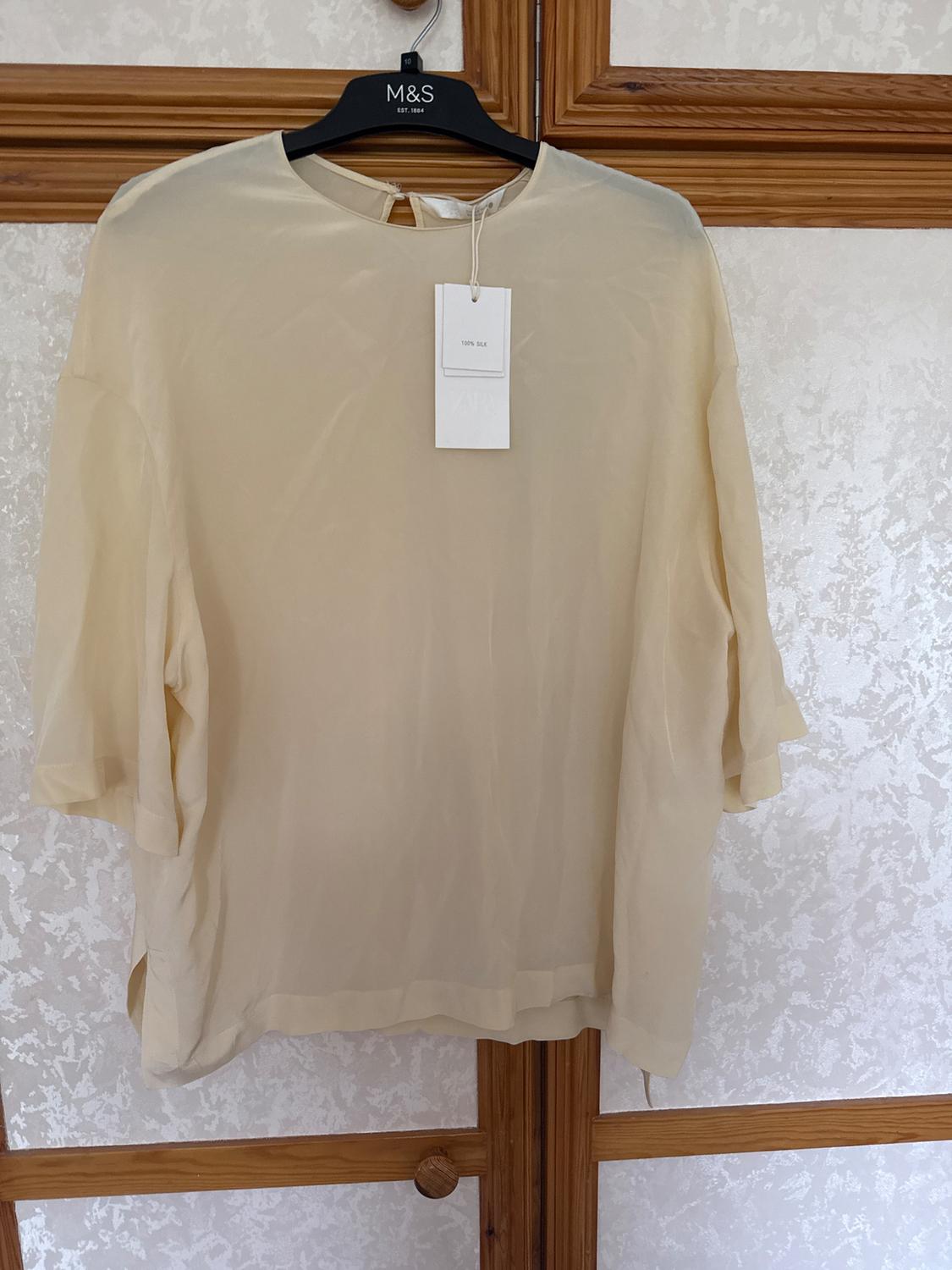 Beige silk blouse hanging on a black hanger with a '100% silk' tag, textured wall background and wooden frame.