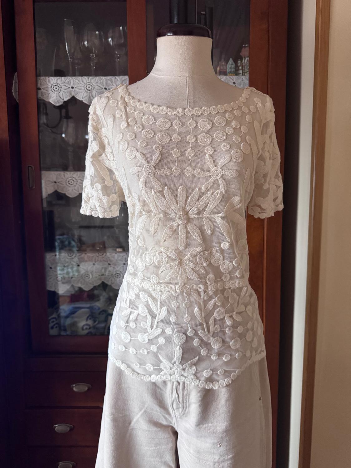 Mannequin in a white blouse embroidered with flowers and light-coloured trousers, in front of a wooden wardrobe with crystal glasses.