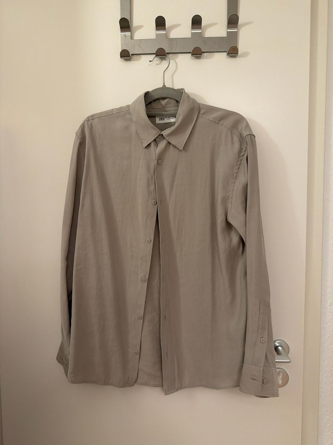 Beige long-sleeved button-up shirt hanging on a hanger on a white door. Label with the text ZARA on the collar.