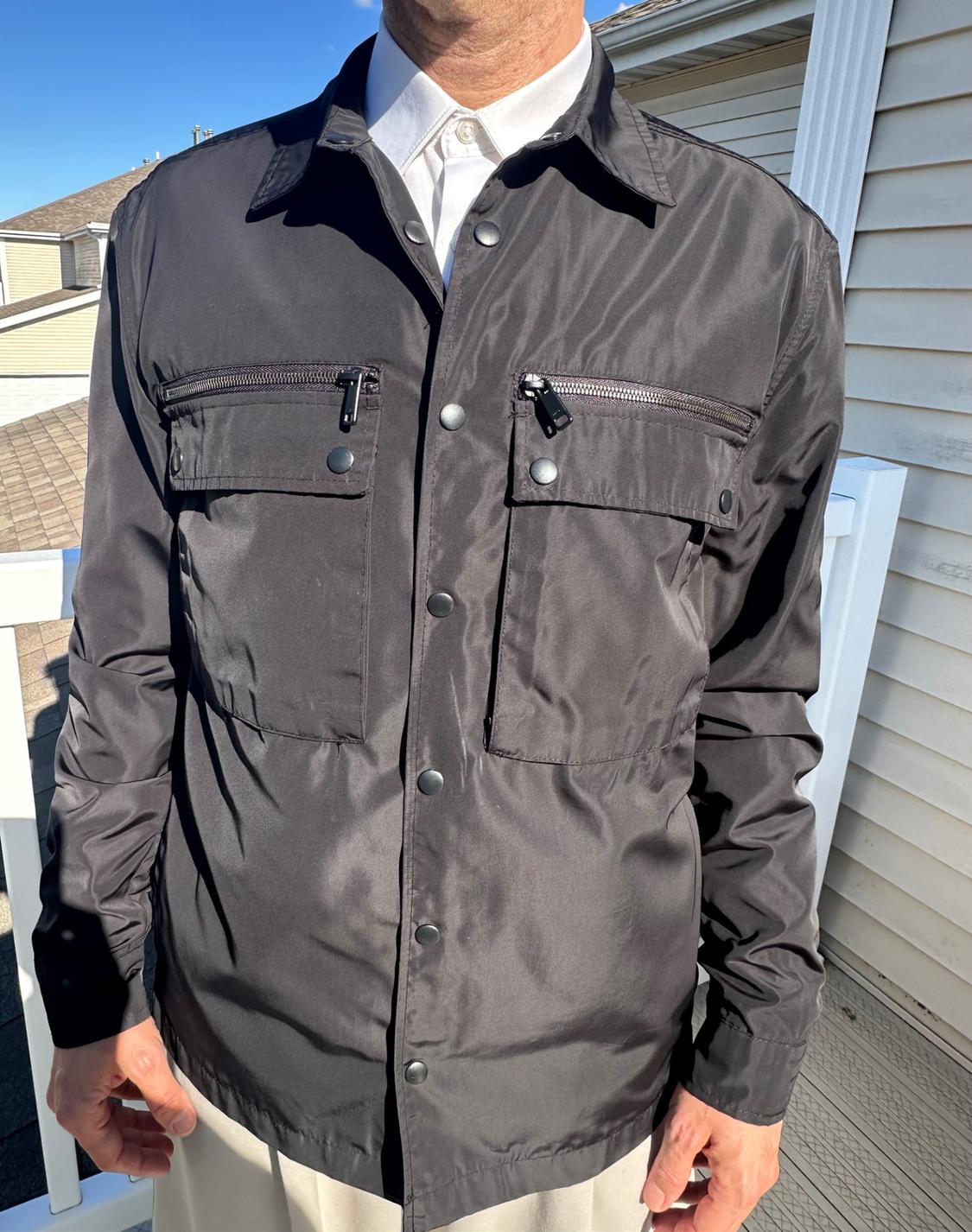 Man in a black button-up and zip-up jacket, white shirt underneath. House and blue sky in the background.