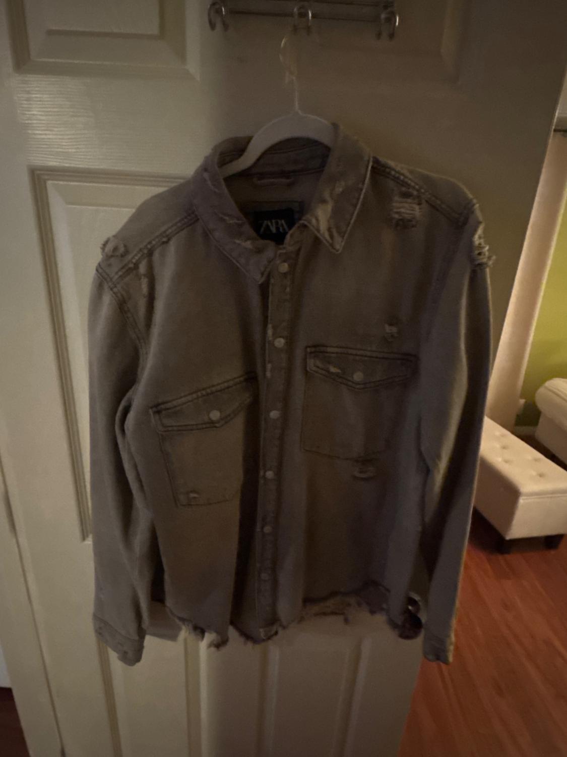 Grey long-sleeve shirt with ripped details and pockets, hanging on a white door. The neck label reads ZARA.
