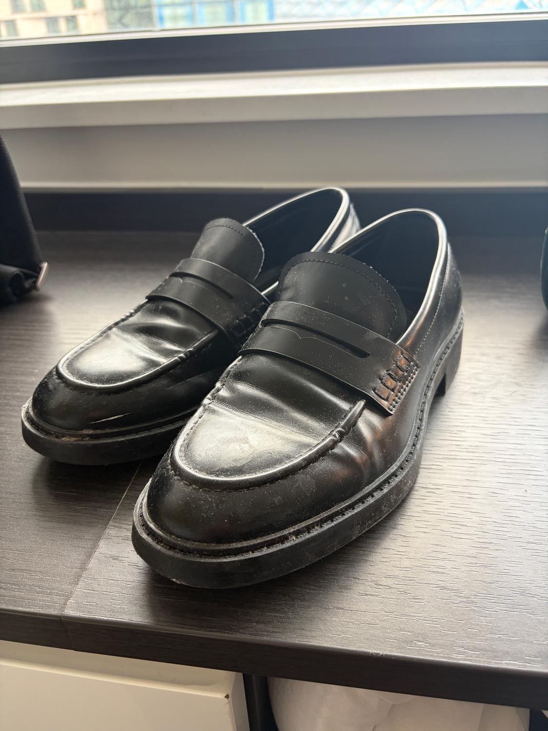 Black leather loafer-style shoes on a dark wooden table, with a window in the background showing buildings.