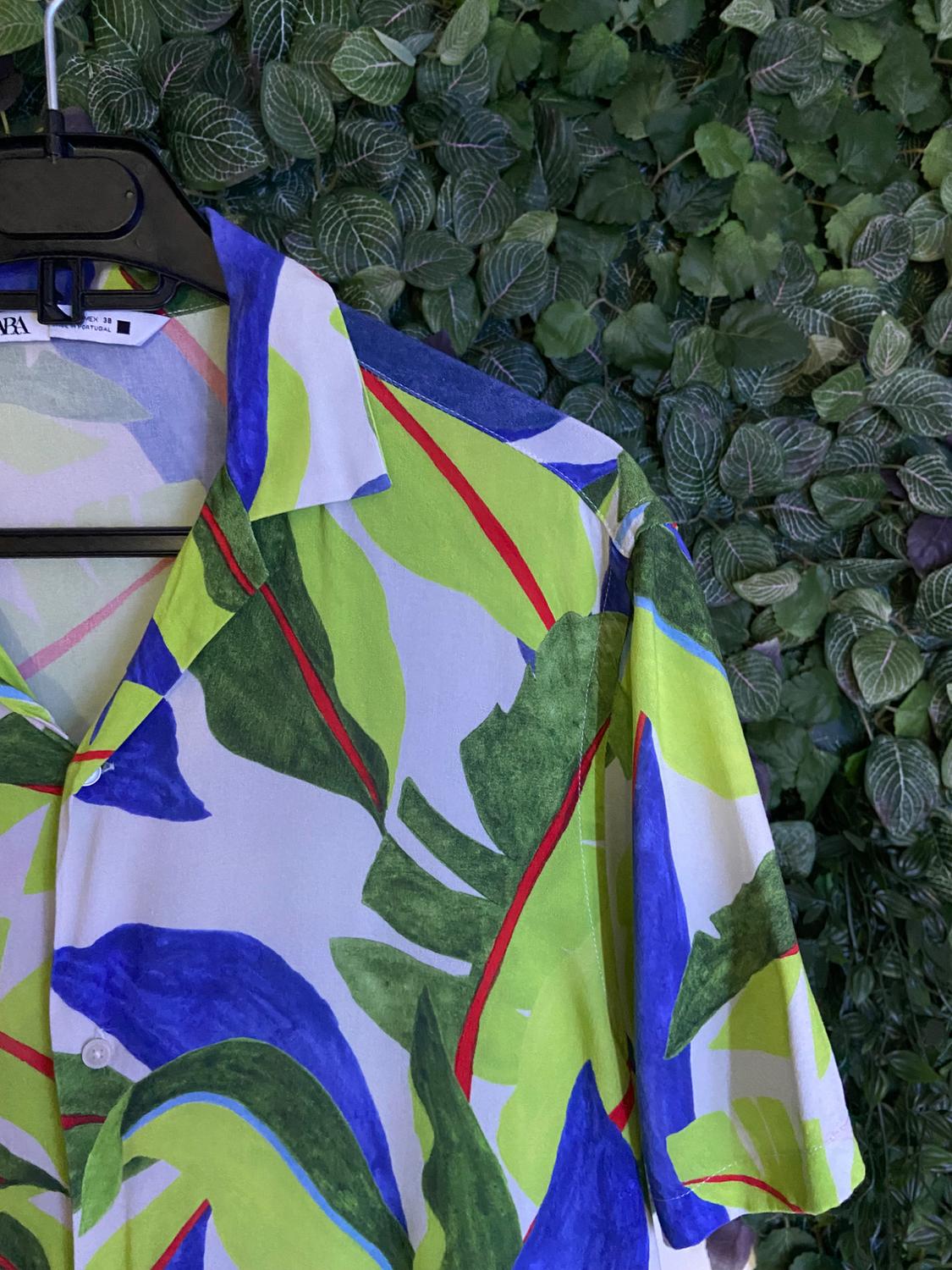 Short-sleeved shirt with a green, blue, and red tropical print, hanging on a black hanger in front of a wall of green leaves.