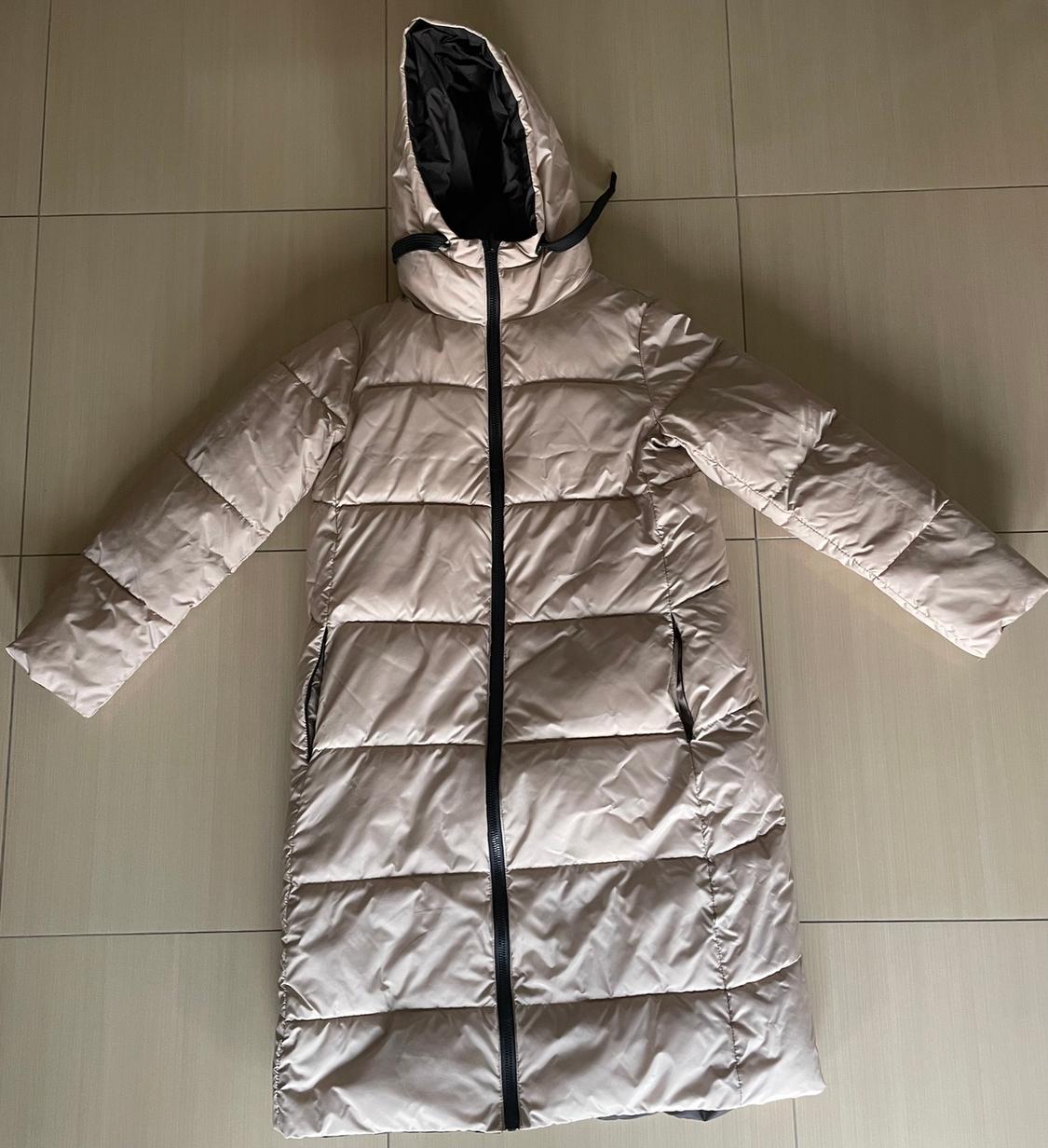 Long beige padded coat with a hood and black zip, laid out on a beige tiled background.