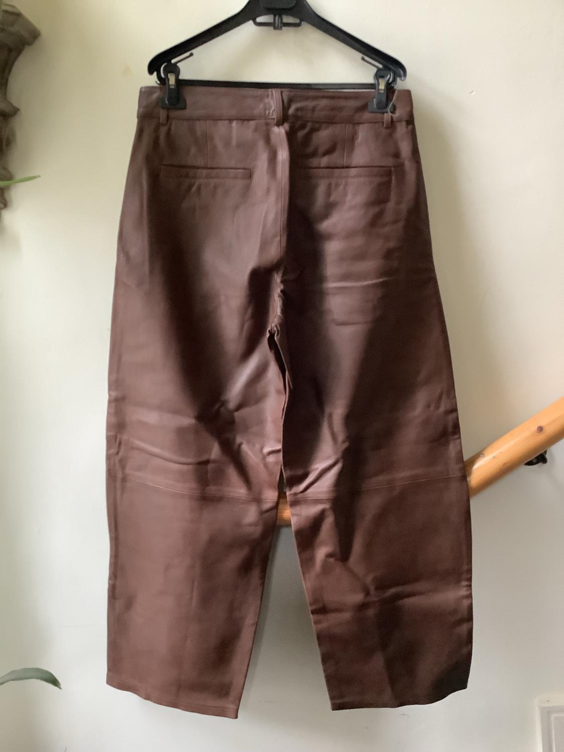 Brown trousers hanging on a black hanger against a white wall. On the right, a wooden handrail can be seen in a casual setting.