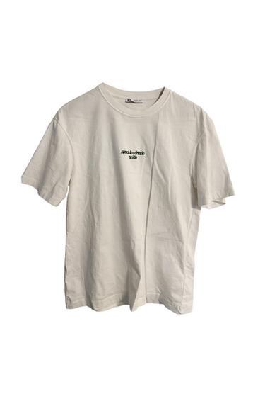 White T-shirt with black text "Necessidade Criado no Rio" on the chest, hung on a white background.