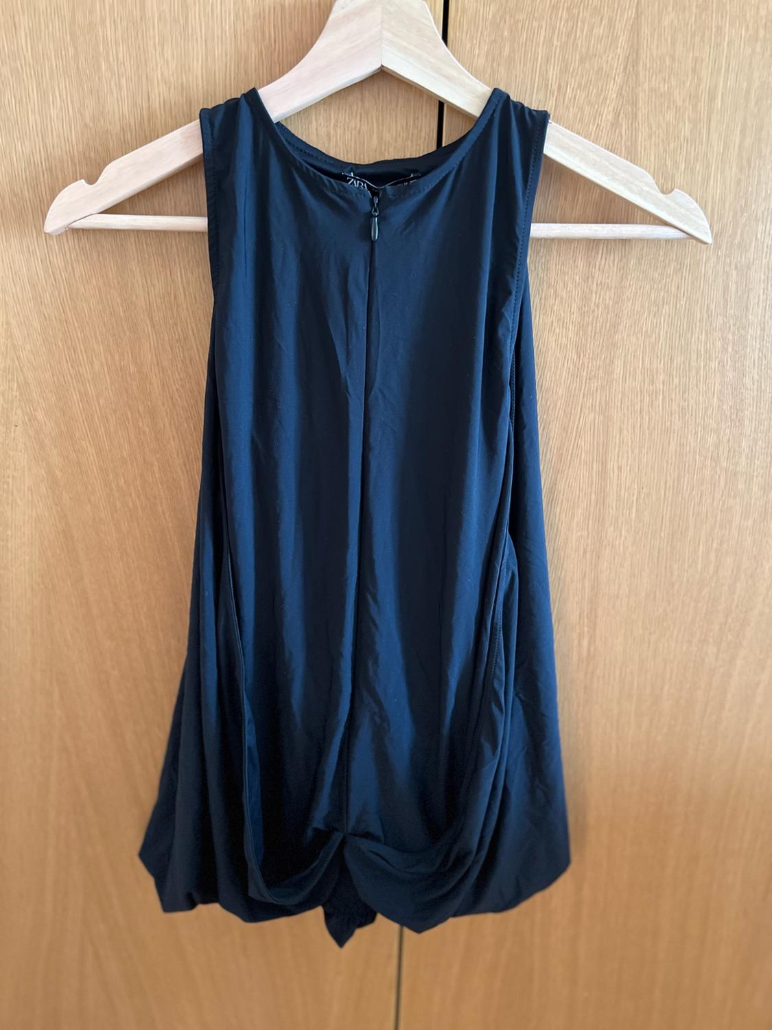 Navy blue sleeveless top with draped hem and zip, hanging on a wooden hanger against a wardrobe background. ZARA label.