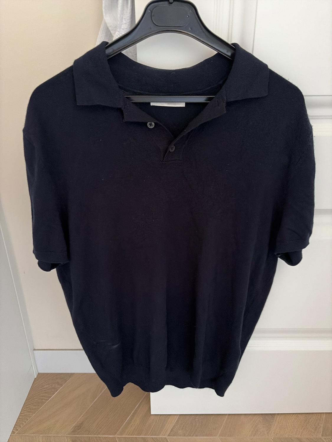 Black short-sleeved polo shirt hanging on a black hanger over a white door. Light wooden floor visible at the bottom.